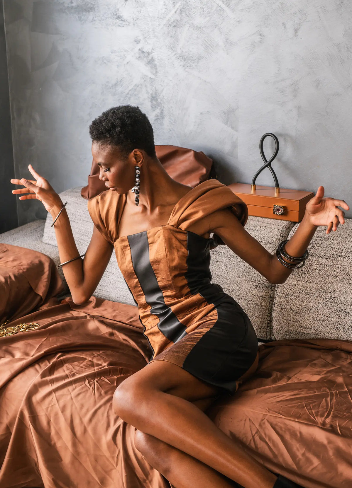 Modèle assise portant une robe courte marron et noire, avec un sac rectangulaire posé derrière elle.